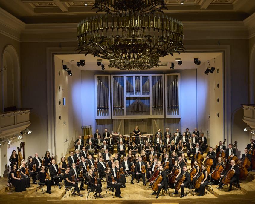 Orkiestra Symfoniczna Filharmonii Śląskiej siedzi na estradzie w składzie około 60 osób, muzycy są ubrani w czarne stroje i trzymają w rękach instrumenty